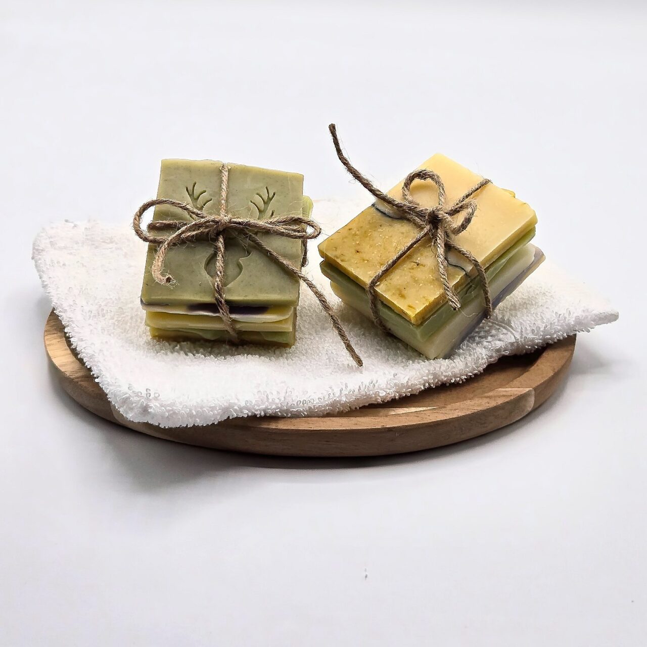 Zwei Sets aus handgemachten Seifenstücken, jeweils mit einer Juteschnur gebunden, liegen auf einem weißen Handtuch auf einem Holztablett. Die Seifen sind in verschiedenen Farben wie Grün, Gelb, Lila und Weiß.